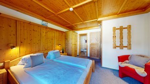 Une pièce aux murs lambrés, avec un lit, un fauteuil rouge et une salle de bain visible à travers une porte ouverte.