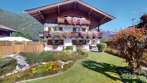 Un chalet traditionnel avec des balcons en bois ornés de fleurs, un petit jardin et un étang.