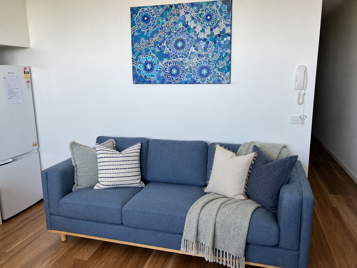 Ein blaues Sofa mit gemusterten Kissen, ein dekorativer blauer Wandbehang, ein weißes Wandtelefon und ein weißer Kühlschrank.
