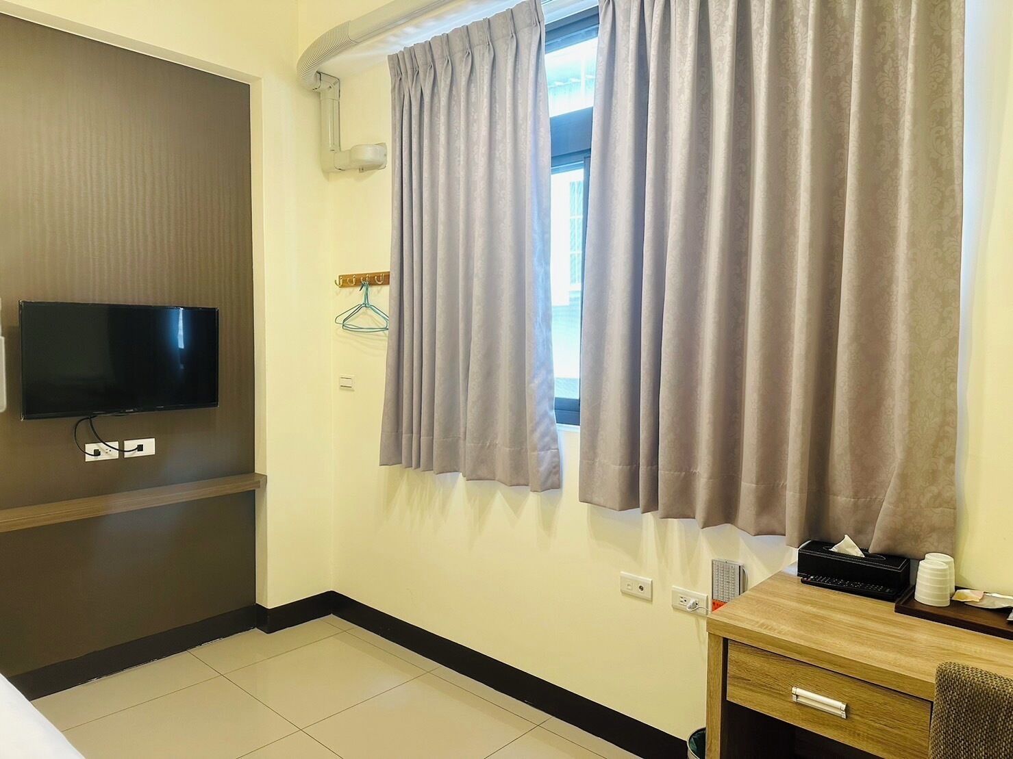 A hotel room with a flat-screen TV mounted on the wall, a wooden desk with a tissue box, and a window with grey curtains.