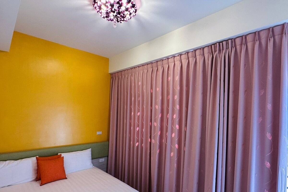 A hotel room with a bed, a yellow wall, and a large window with curtains.