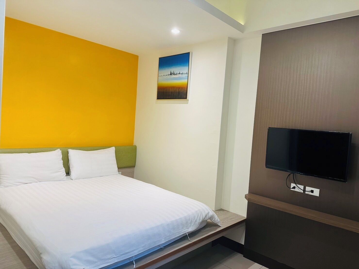 Una habitación de hotel con una cama, un televisor montado en la pared y un cuadro sobre la cama.