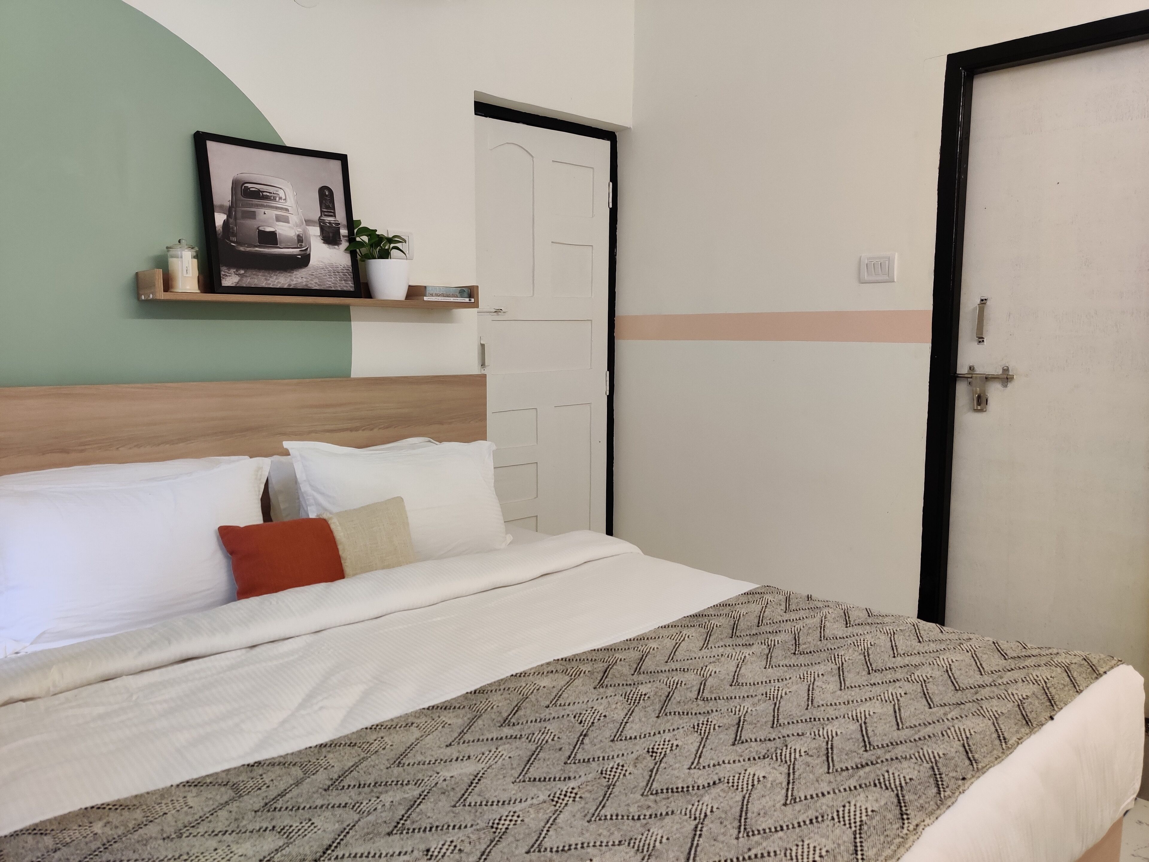 A neatly made bed with white and patterned linens, a headboard, and a bedside shelf with a plant and a framed photo.