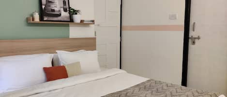 Una cama bien hecha con ropa de cama blanca y estampada, un cabecero y un estante junto a la cama con una planta y una foto enmarcada.