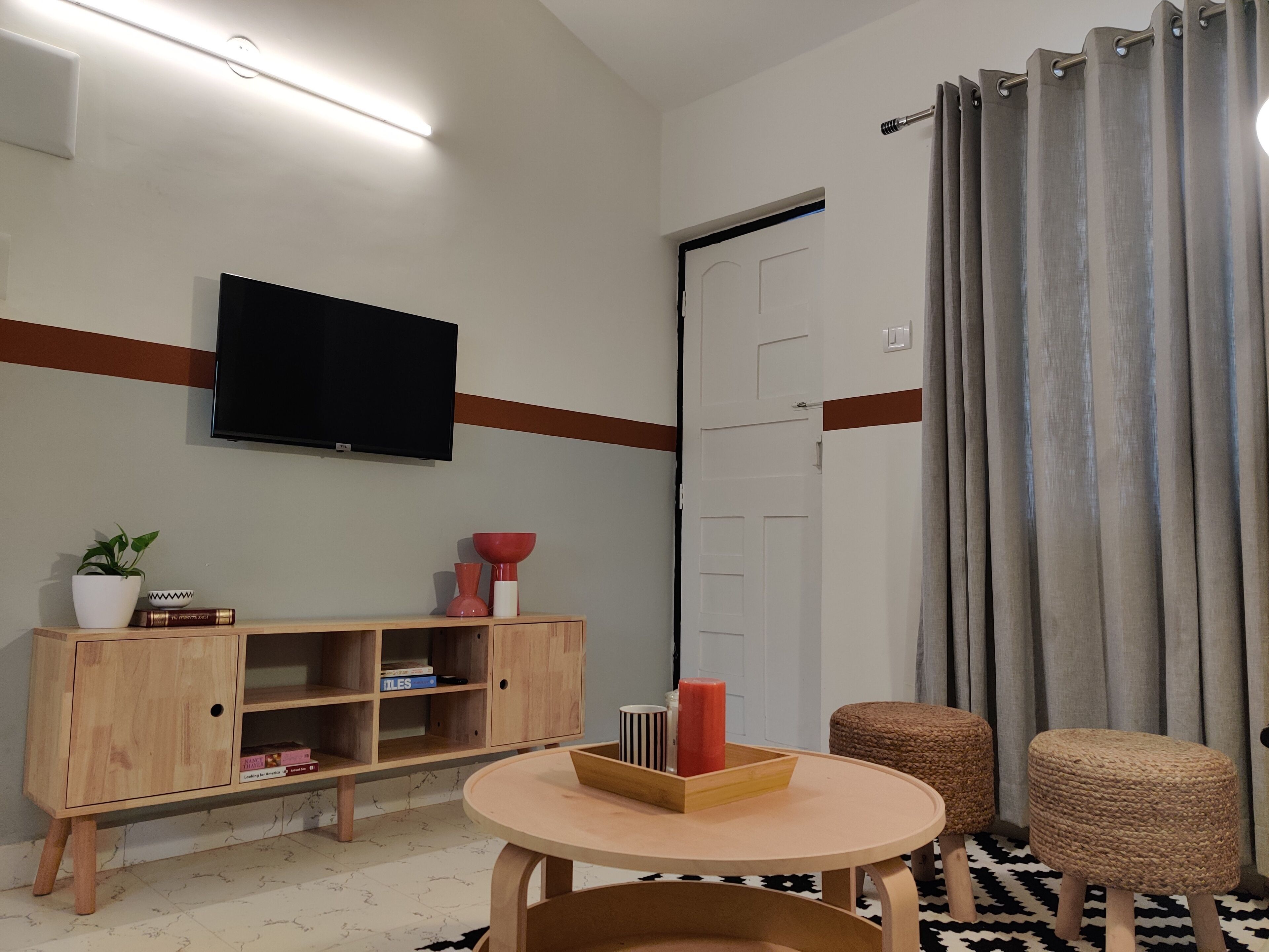 Een moderne woonkamer met een flatscreen televisie aan de muur, een houten kast met boeken, een klein rond tafeltje met een dienblad, en een grijze gordijn over een schuifdeur.