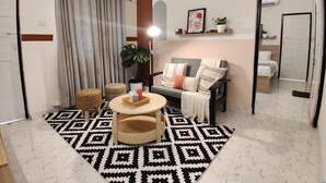 A living room with a sofa, a round coffee table, a patterned rug, a potted plant, and a wall-mounted shelf with books and a picture frame. - Olive Hotel Calangute Niwasa by Embassy Group (Calangute)