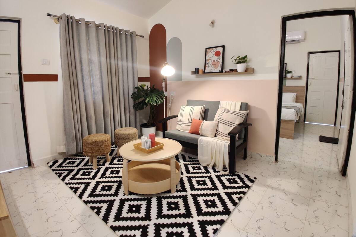 Ein Wohnzimmer mit einer Couch, einem runden Couchtisch, einem gemusterten Teppich, einer Topfpflanze und einem an der Wand befestigten Regal mit Büchern und einem Bilderrahmen.