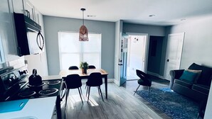 A modern kitchen with a dining area, a living room with a sofa and a rug, and a hallway leading to another room.