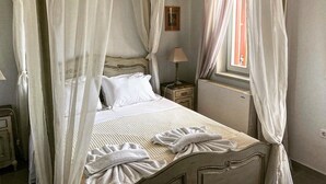 Ein Bett mit Baldachin, ausgestattet mit weißen Leinen und dekorativen Kissen, ein Nachttisch mit Lampe und ein Fenster mit Vorhängen.