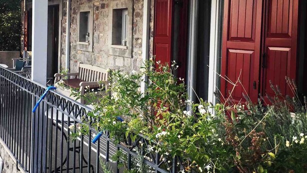 Ein Steinhaus mit roten Fensterläden, einer überdachten Terrasse und einem Metallzaun.