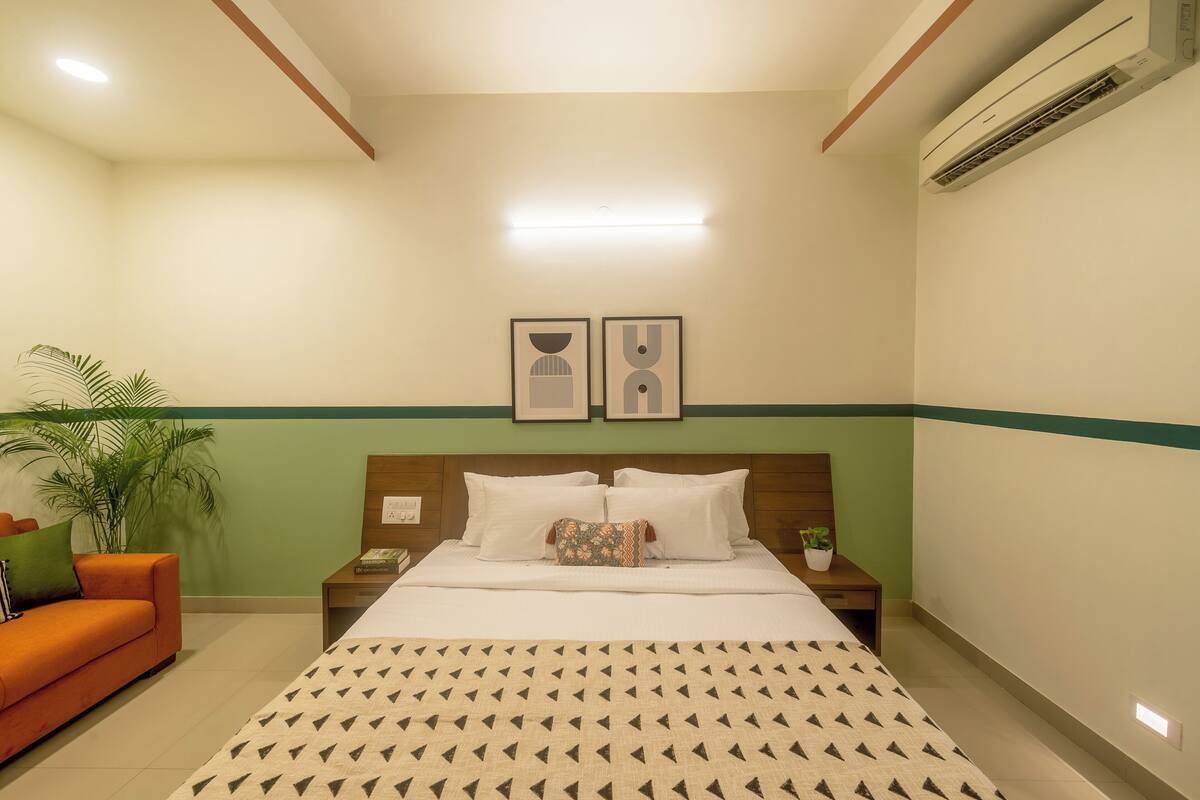 A hotel room with a bed, a nightstand, a bedside table, a lamp, a potted plant, and a sofa.