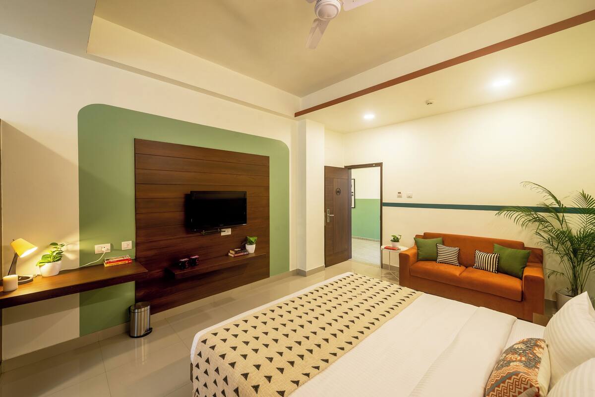 A modern hotel room with a large bed, a TV mounted on a green wall, a wooden desk, and an orange sofa with pillows.