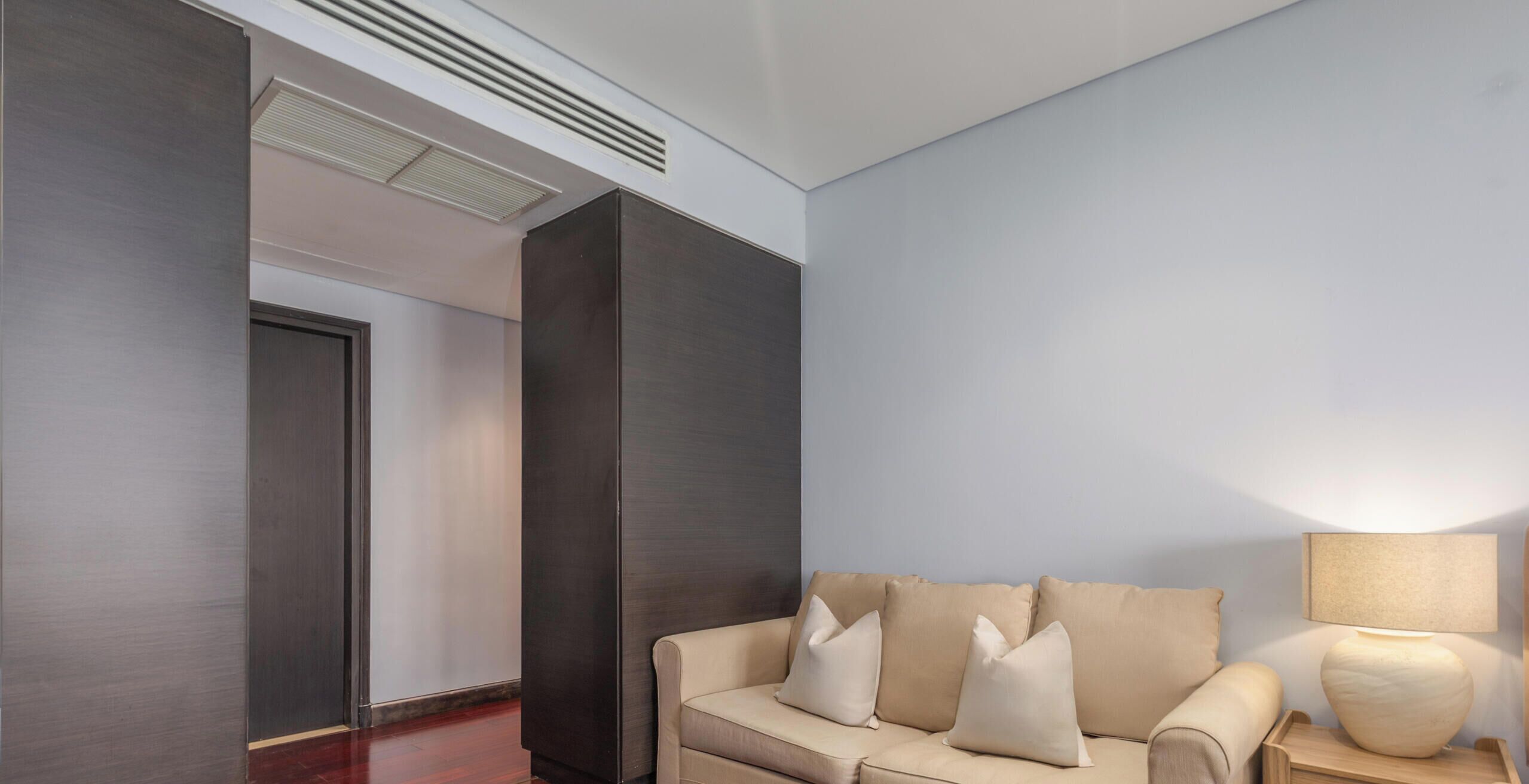 A modern hotel room with a beige sofa, a wooden nightstand with a lamp, and a dark-colored wardrobe.
