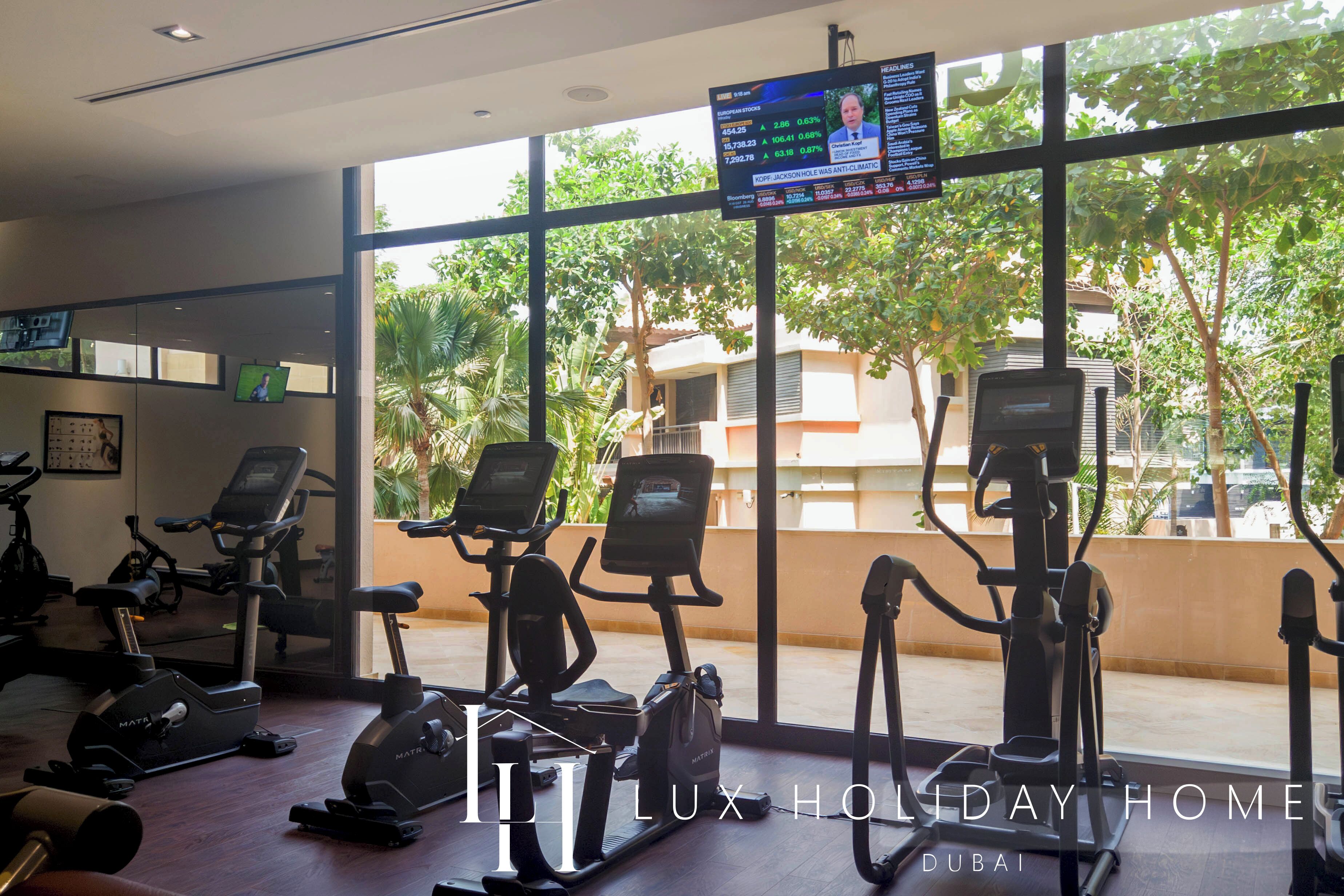 Ein Fitnessstudio mit Ergometer und Cardiogeräten, großen Fenstern mit Blick auf Grünflächen und Gebäude, sowie einem Fernsehbildschirm, der einen Nachrichtensender zeigt.
