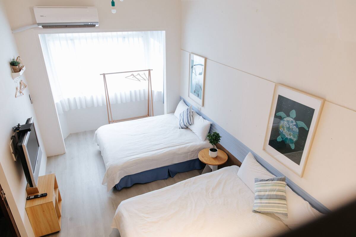 Quarto de hotel com duas camas, uma televisão, uma mesinha com uma planta e obras de arte nas paredes.