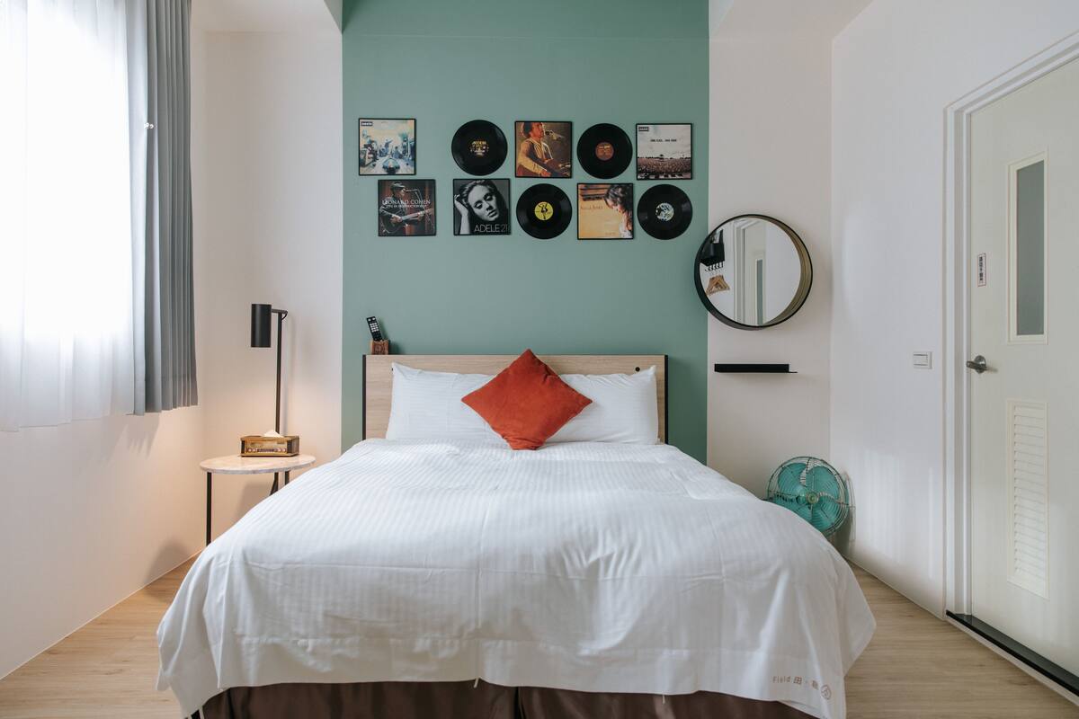Um quarto bem organizado com uma cama, uma coleção de discos na parede, uma mesa de cabeceira com abajur e uma porta.