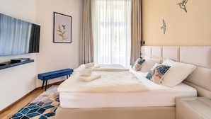 A hotel room with two beds, a flat-screen TV, a bench, and a large window with sheer curtains. - Luxury Residence Kmochova (Prague)