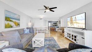 A modern living room with a grey sofa, a white coffee table, and a flat-screen TV. There is a dining area with white chairs and a kitchen in the background. - Beautiful Gulffront, Beachfront Phoenix VI Condo!! (Orange Beach)