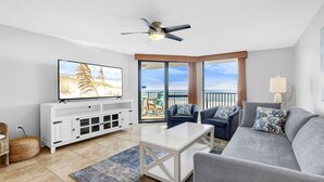 A living room with a flat-screen TV, white entertainment unit, grey sofa, and a balcony with a view of the beach. - Beautiful Gulffront, Beachfront Phoenix VI Condo!! (Orange Beach)