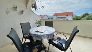 Una mesa redonda con mantel estampado, cuatro sillas, un adorno con forma de corazón y plantas en macetas en un balcón con vistas a edificios y un cielo nublado.
