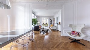 Um quarto amplo com uma mesa de jantar de vidro, cadeiras transparentes, uma poltrona branca estofada com uma almofada colorida e piso de madeira em padrão herringbone.