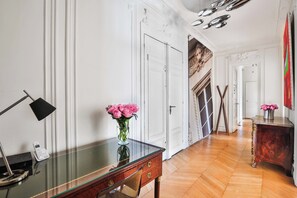 Une pièce comprenant un bureau avec un plateau en verre, un vase de fleurs, un téléphone et une table d’appoint en bois sur laquelle se trouve un autre vase de fleurs.