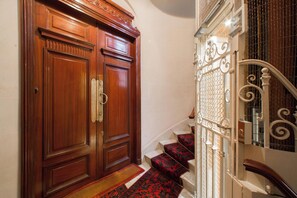Une porte à deux battants en bois avec une poignée en laiton, un escalier recouvert d’une moquette rouge avec des rampes blanches, et un mur blanc.