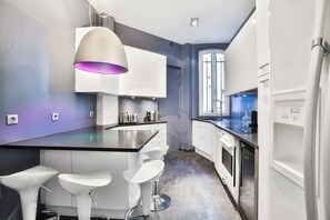 Une cuisine moderne avec des armoires blanches, un comptoir noir et un luminaire suspendu violet. On y trouve trois tabourets de bar blancs et une fenêtre donnant sur la ville.