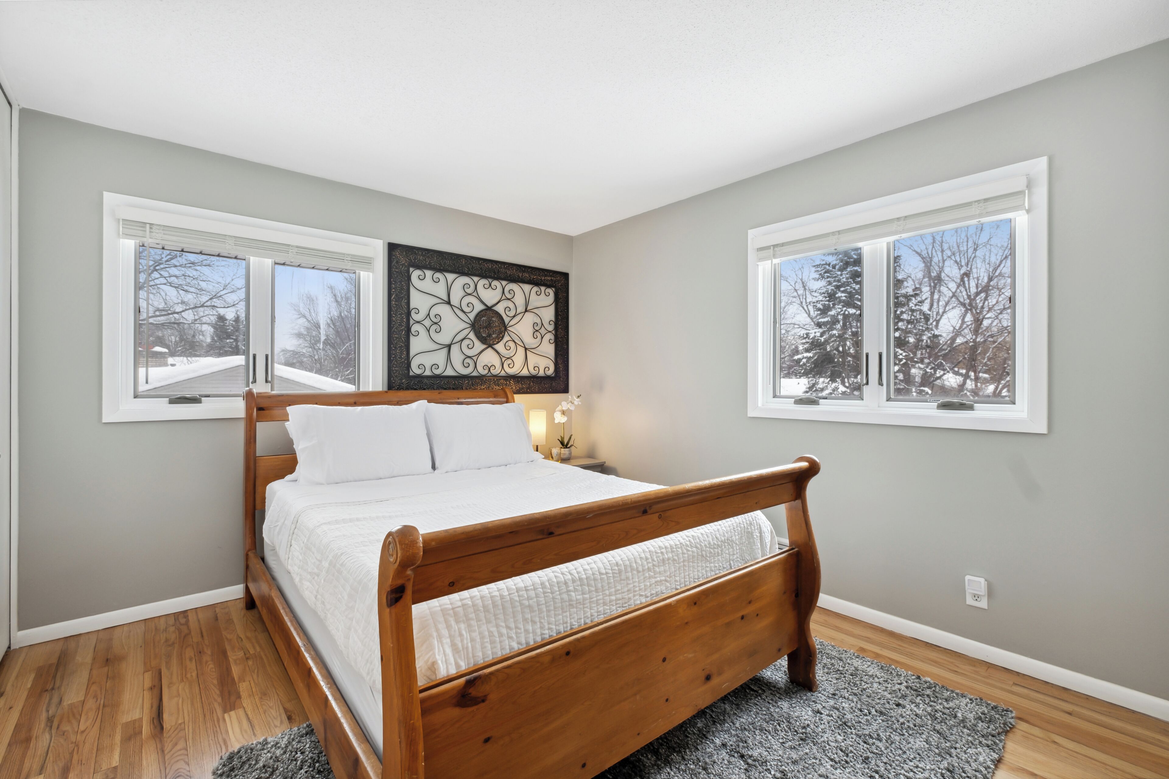 A bedroom with a wooden bed, two windows, a wall art piece, and a nightstand.