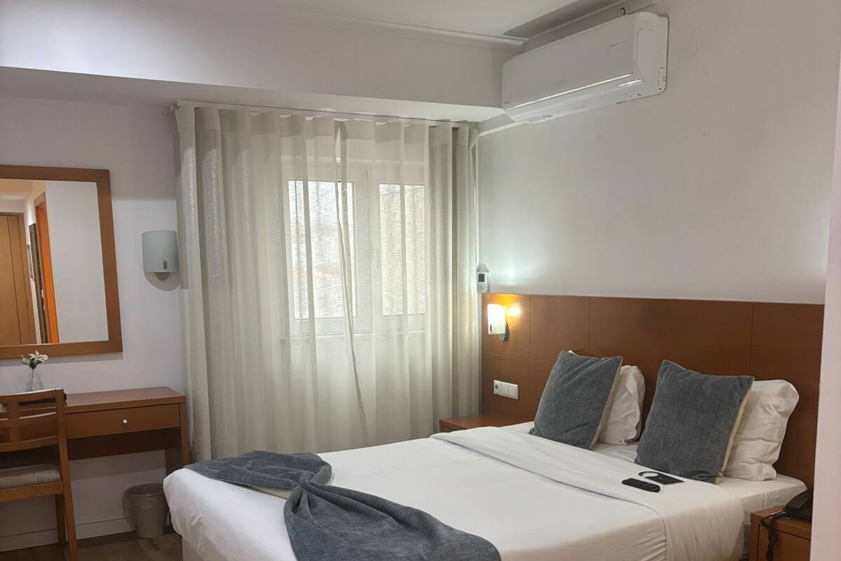 A hotel room with a large bed, a wooden desk, a mirror, and a window with curtains.