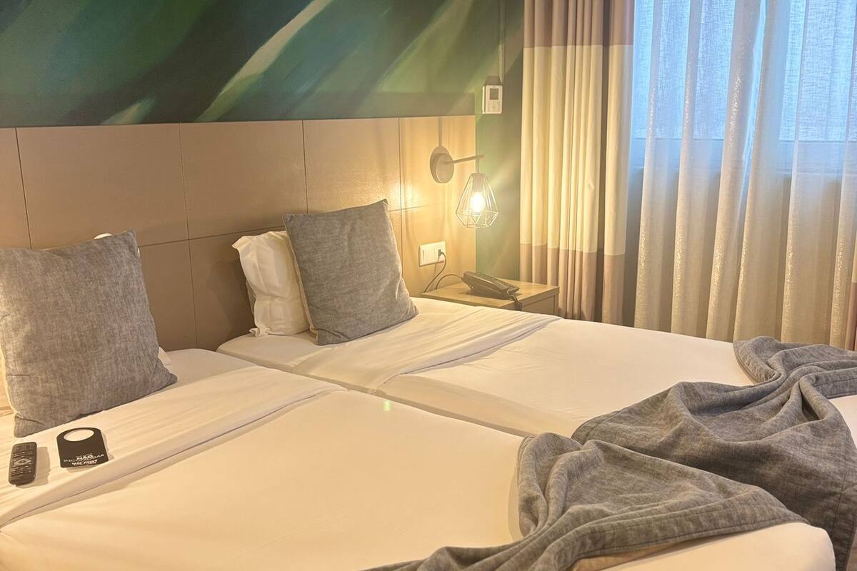 A hotel room with two beds, a wall-mounted air conditioner, a bedside lamp, and a window with curtains.