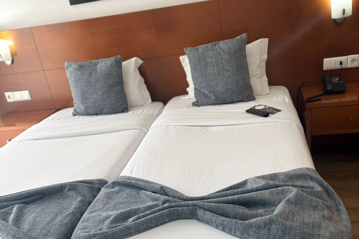 A double bed with white linens and gray throw pillows, a framed floral artwork above, and a bedside table with a phone and lamp.