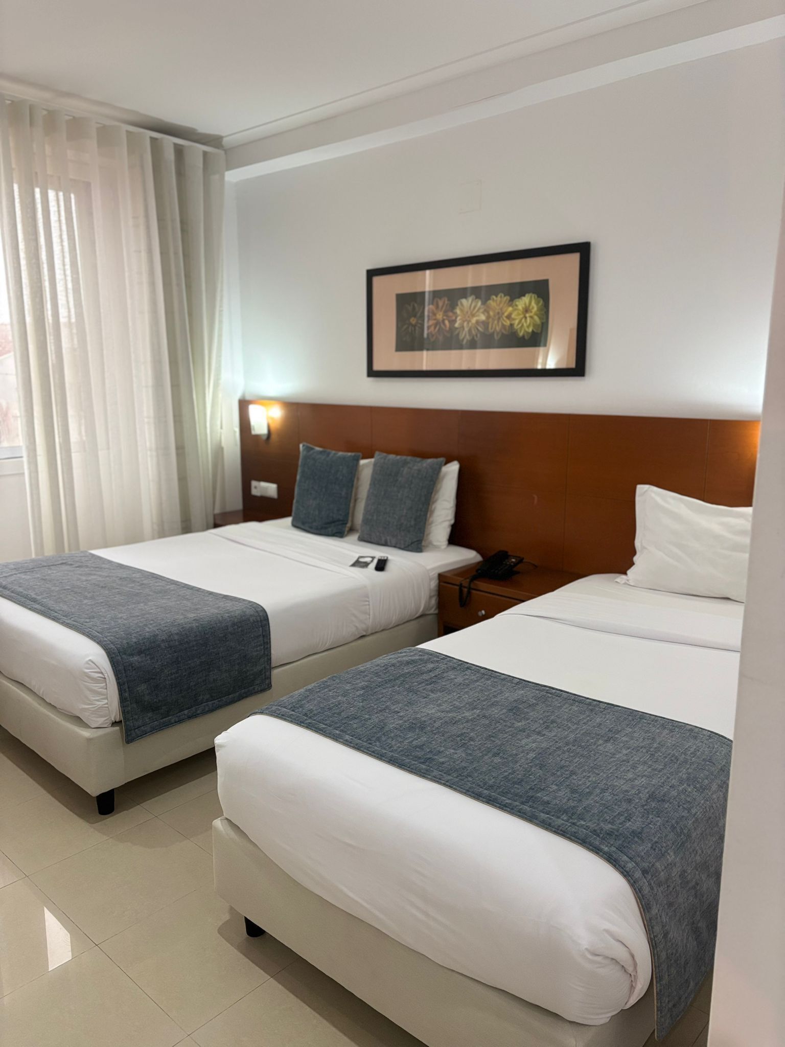 A hotel room with two beds, a framed floral artwork on the wall, and a nightstand with a phone.