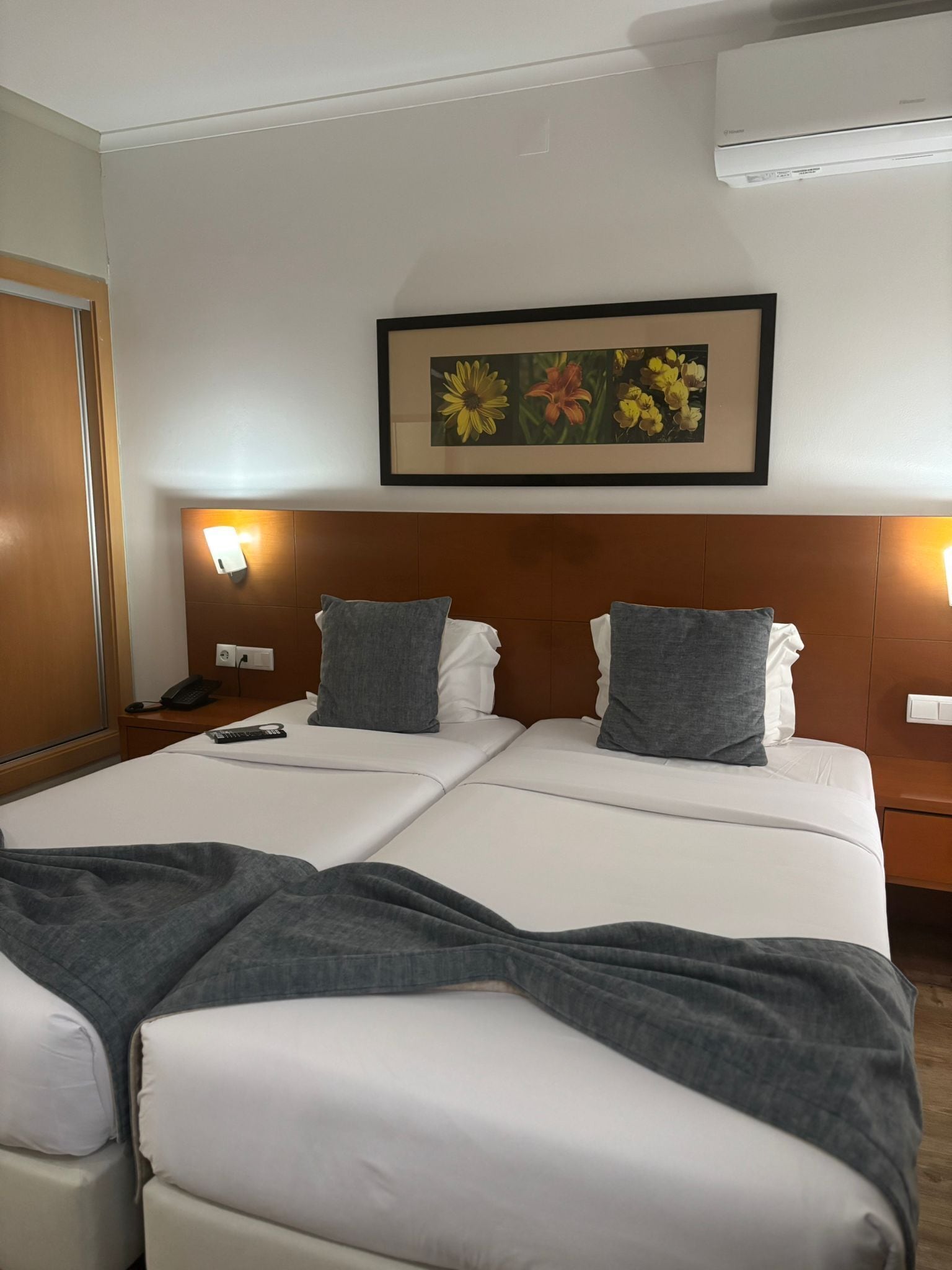 A hotel room with a double bed, two pillows, a bedside table with a phone, a framed floral artwork above the bed, and an air conditioning unit on the wall.