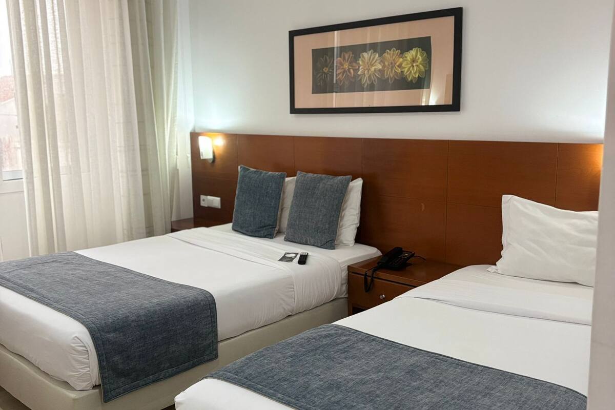 A hotel room with two beds, a framed floral artwork on the wall, and a bedside table with a lamp.