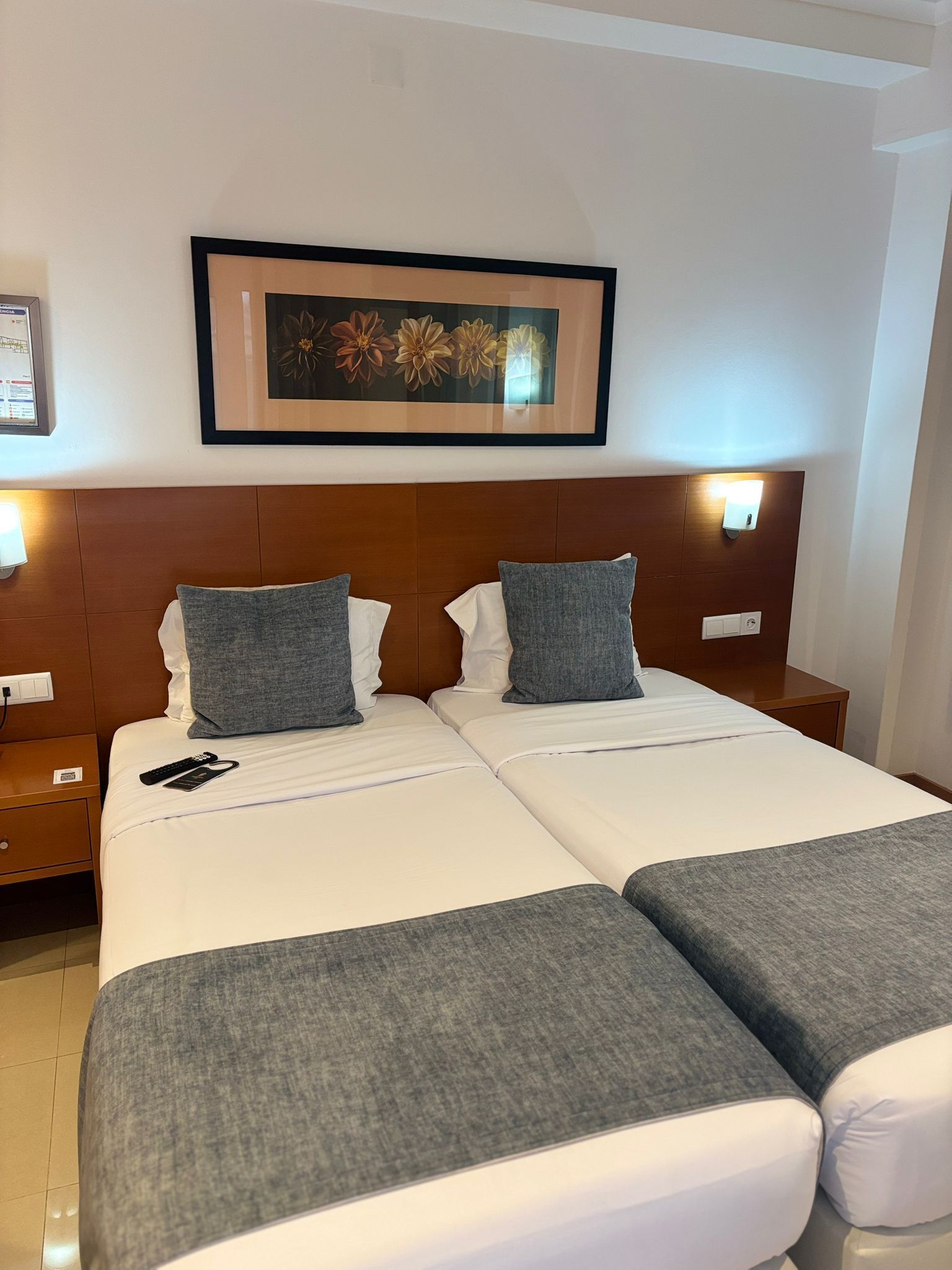 A hotel room with two beds, a wooden headboard, a framed floral artwork, and a nightstand with a lamp.