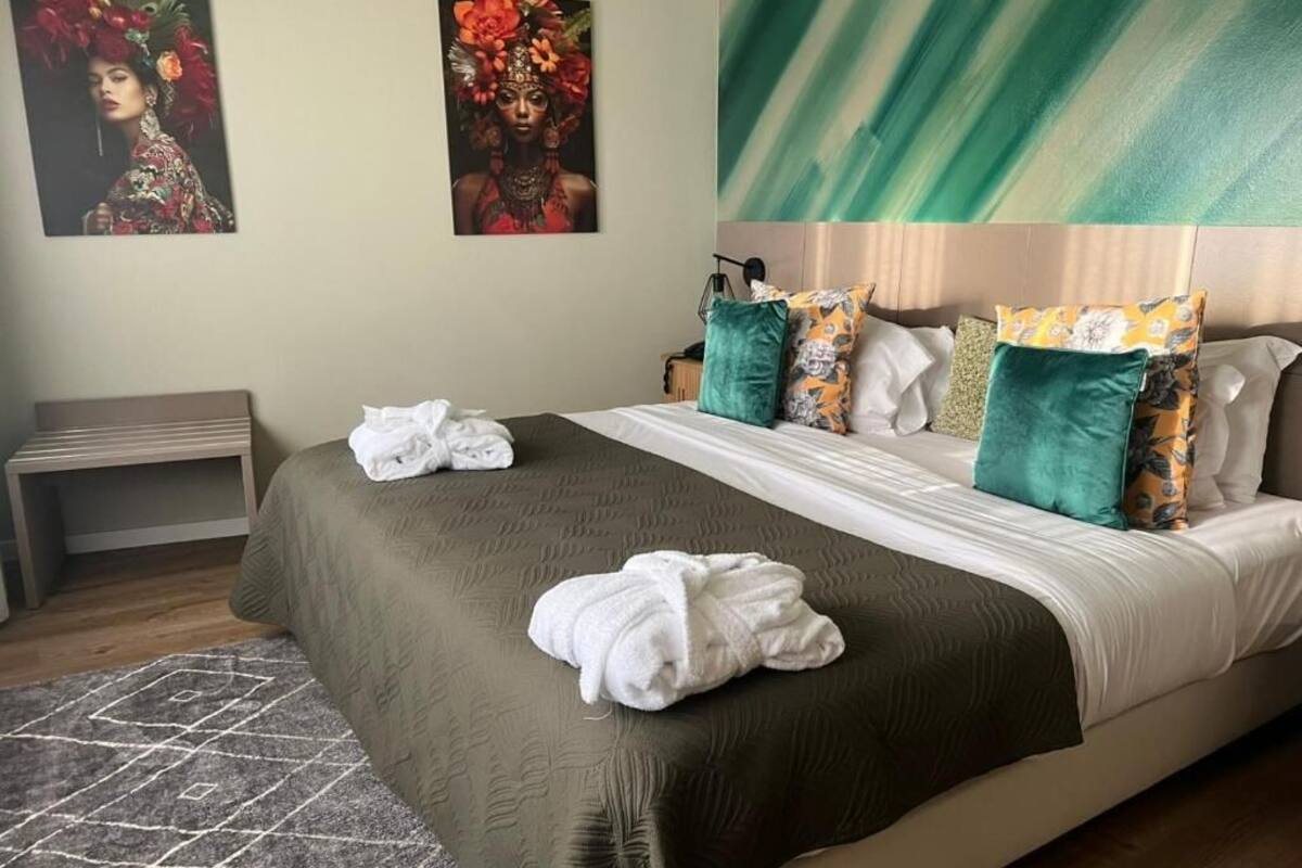 A neatly made bed with a green bedspread, white pillows, and a headboard with patterned cushions. There are two paintings on the wall above the bed.
