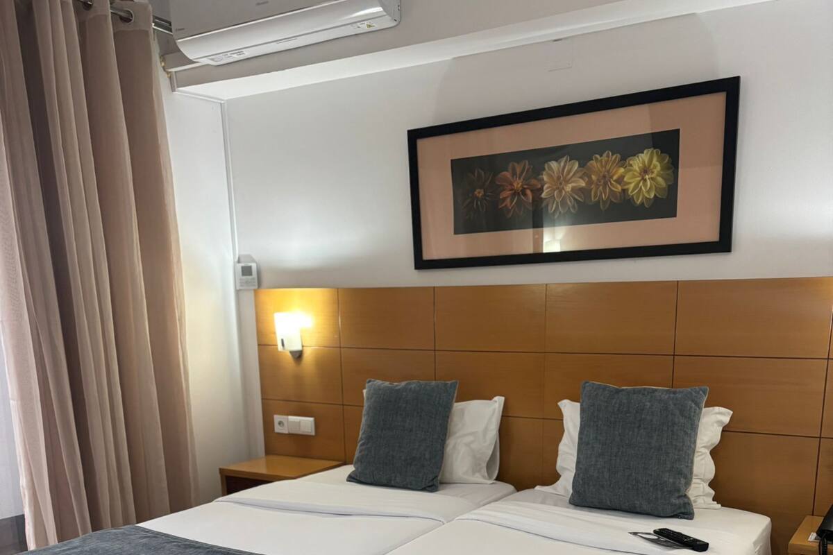 A double bed with a wooden headboard, two bedside tables, a framed floral artwork, and a wall-mounted air conditioner.