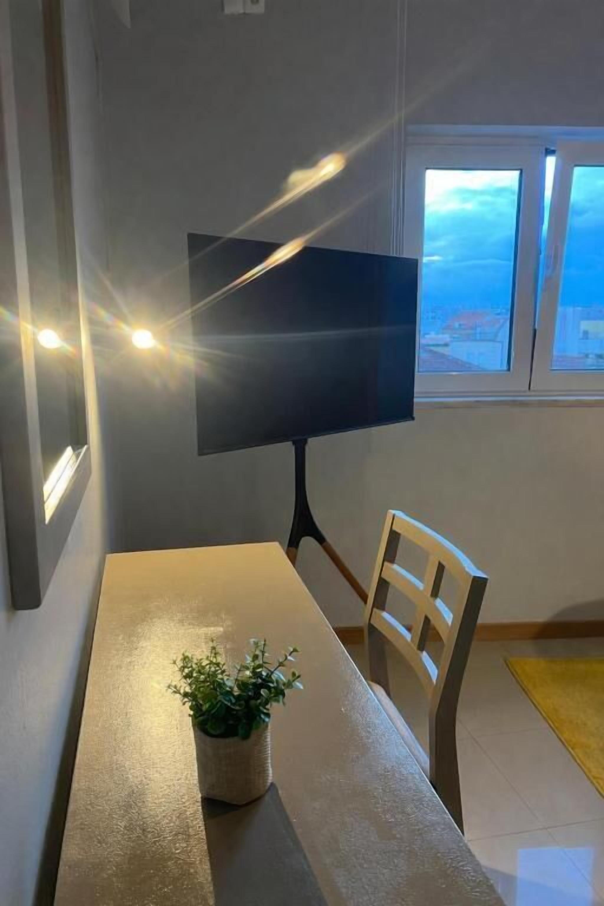 A room with a large flat-screen TV mounted on the wall, a dining table with a potted plant, and a view through a window.