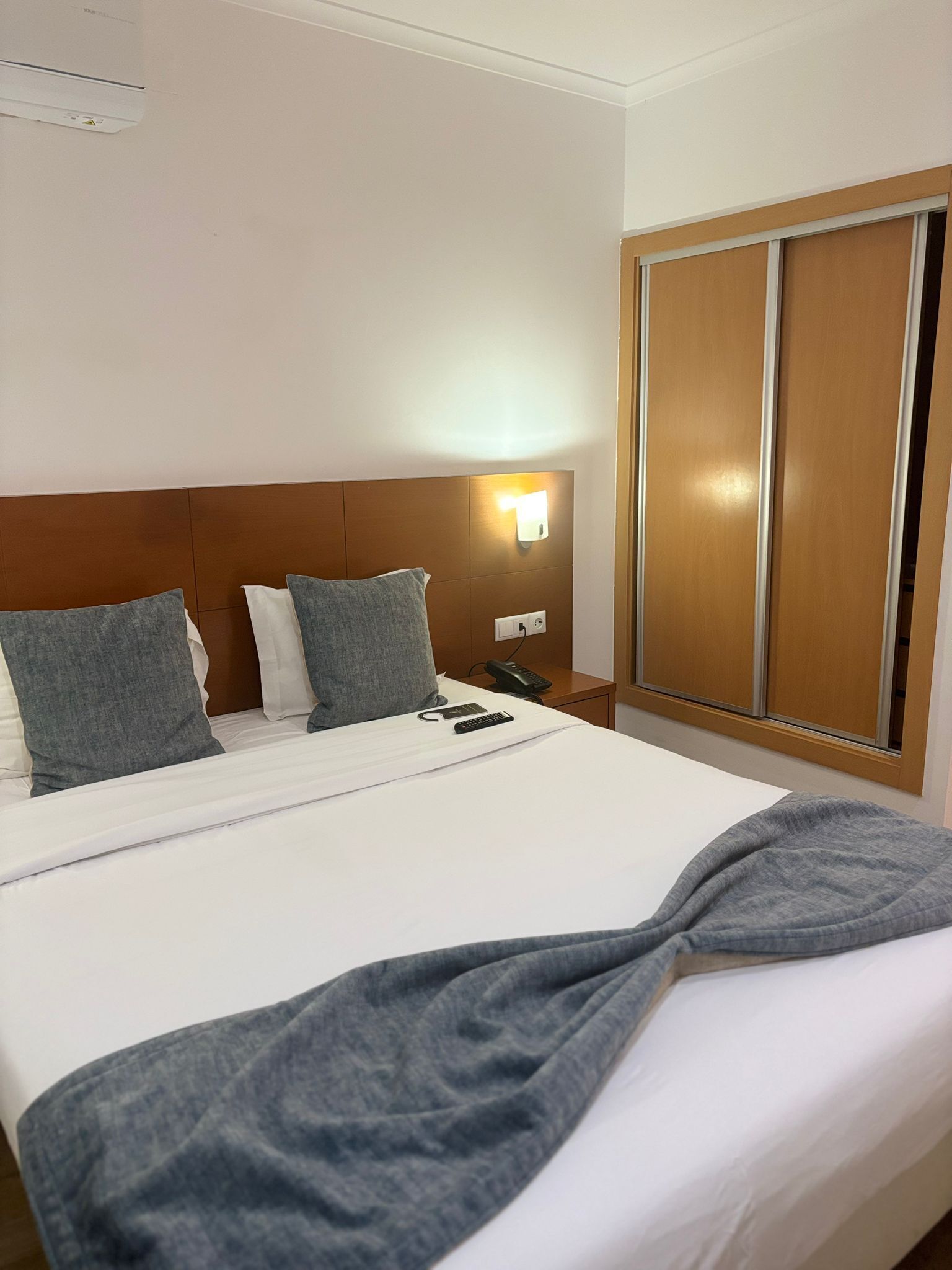 A hotel room with a large bed, a wooden headboard, a bedside table with a phone and remote controls, and a wardrobe with sliding doors.