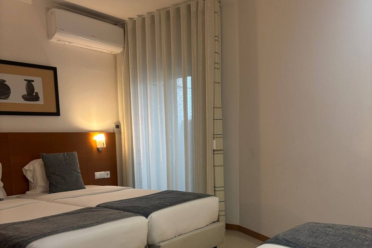 A hotel room with a bed, a sofa bed, a bedside table with a lamp, a framed picture on the wall, and a window with curtains.