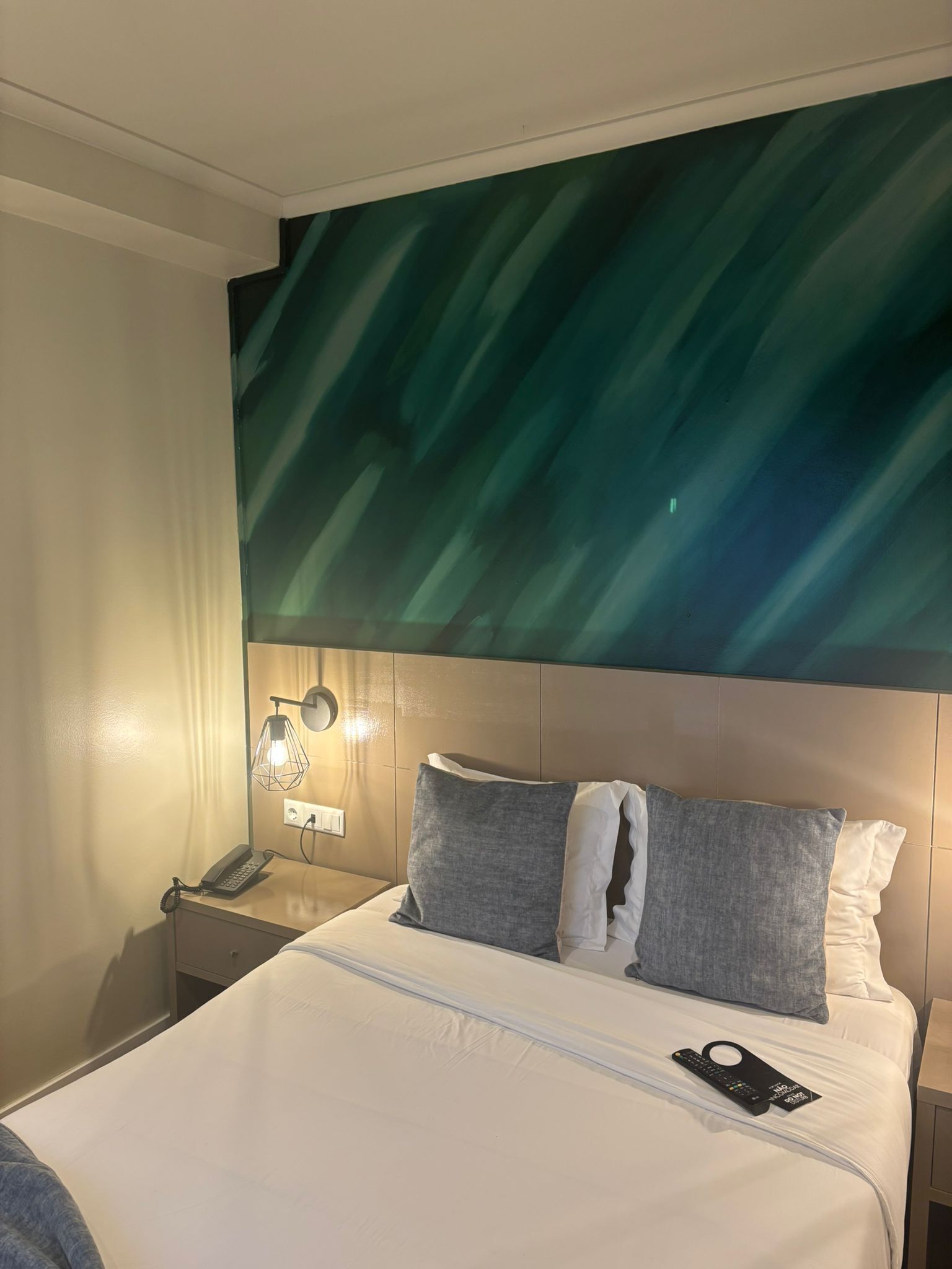 A hotel room with a bed, a nightstand with a lamp, and a wall with a green abstract design.