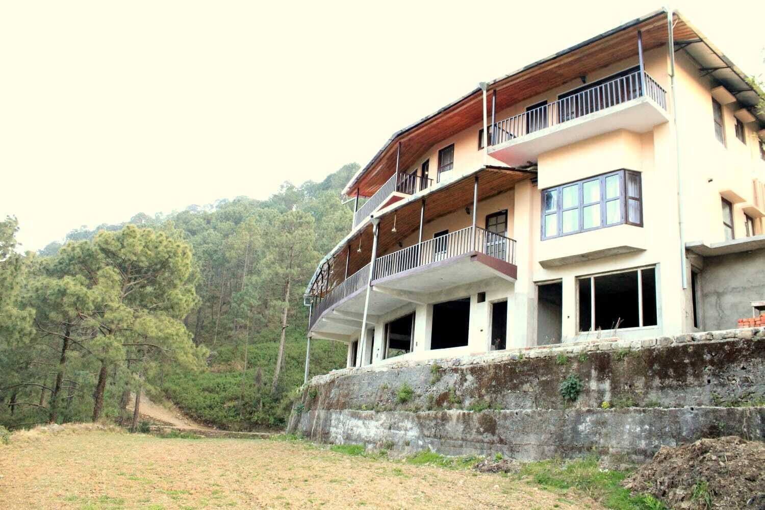 Un edificio de varios pisos con balcones, un cimiento de hormigón y un paisaje montañoso cubierto de bosque al fondo.
