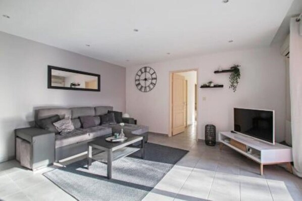 Un salon moderne avec un canapé gris, une table basse, un téléviseur posé sur un meuble et une horloge au mur.