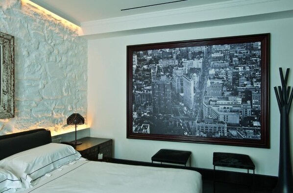 A bedroom with a cityscape view, a stone wall, a bed with white linens, a nightstand, and a lamp.