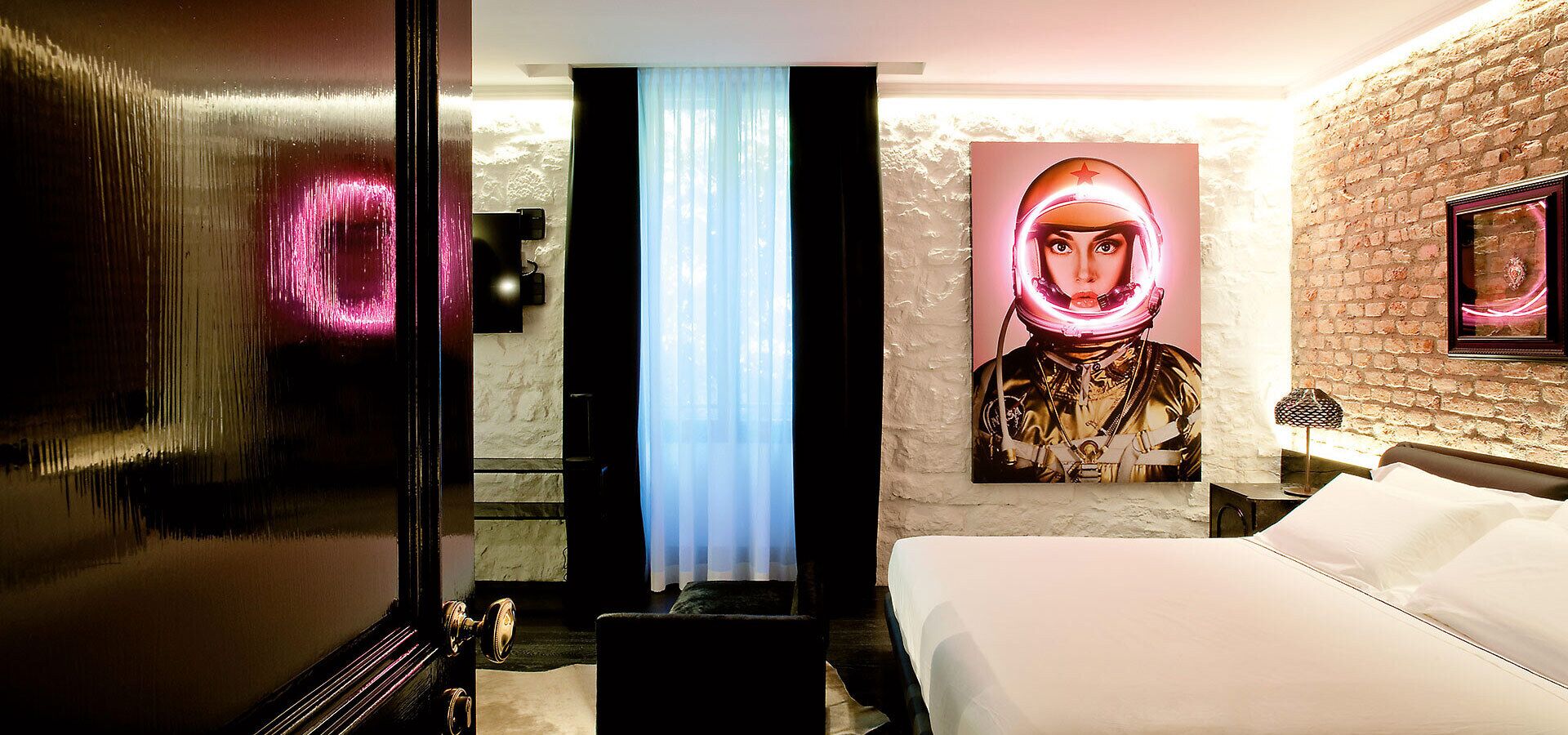 Ein Hotelzimmer mit einem Bett, einem großen Bild eines Raumanzugs, einer Backsteinwand und einer Tür mit spiegelnder Oberfläche.