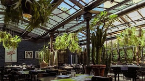 A restaurant with a high ceiling and glass panels, featuring hanging plants and a rustic stone wall. Tables are set with menus and glasses. - Areadocks Boutique Hotel (Brescia)