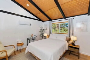A bedroom with a wooden ceiling, a bed with white bedding, a nightstand with a lamp, a small table with a lamp, a chair, and a window with curtains. - Eagle Peak Walk to Village & Lake Fire Pit (Big Bear Lake)