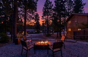 Una cabaña con fogata y asientos exteriores, rodeada de árboles y vistas al atardecer.