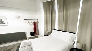 A hotel room with a large bed, a bedside table, a nightstand, a radiator, a window with curtains, and a wall-mounted towel rack. - Erin Court Mansion -Suite 11 (South Croydon)